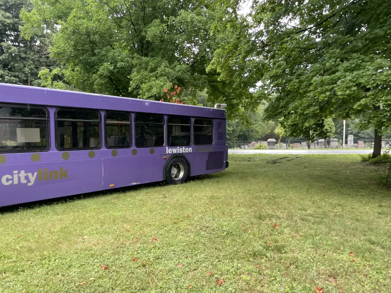 Rear half of the purple bus resting on grass under maple trees, wheels sunk into the lawn