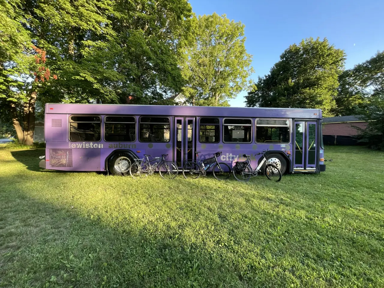The purple bus in full side profile on the lawn in evening light, four bicycles leaning against it