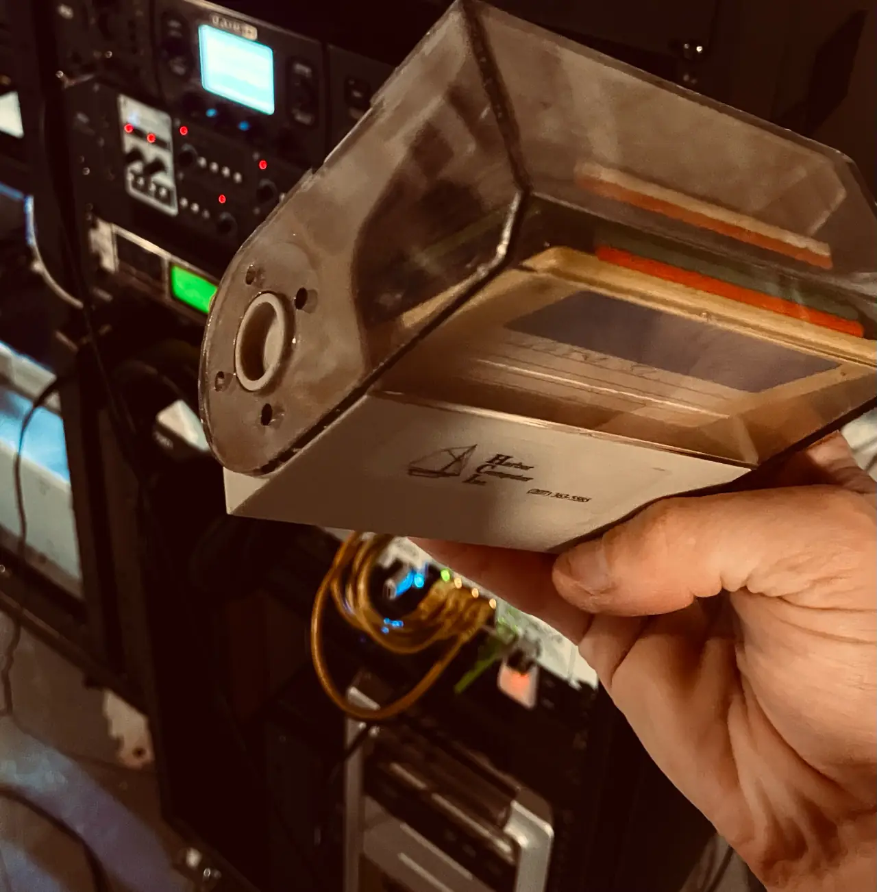 A hand holding up a palm-sized electronic device in a clear acrylic case in front of a rack of audio gear with lit displays