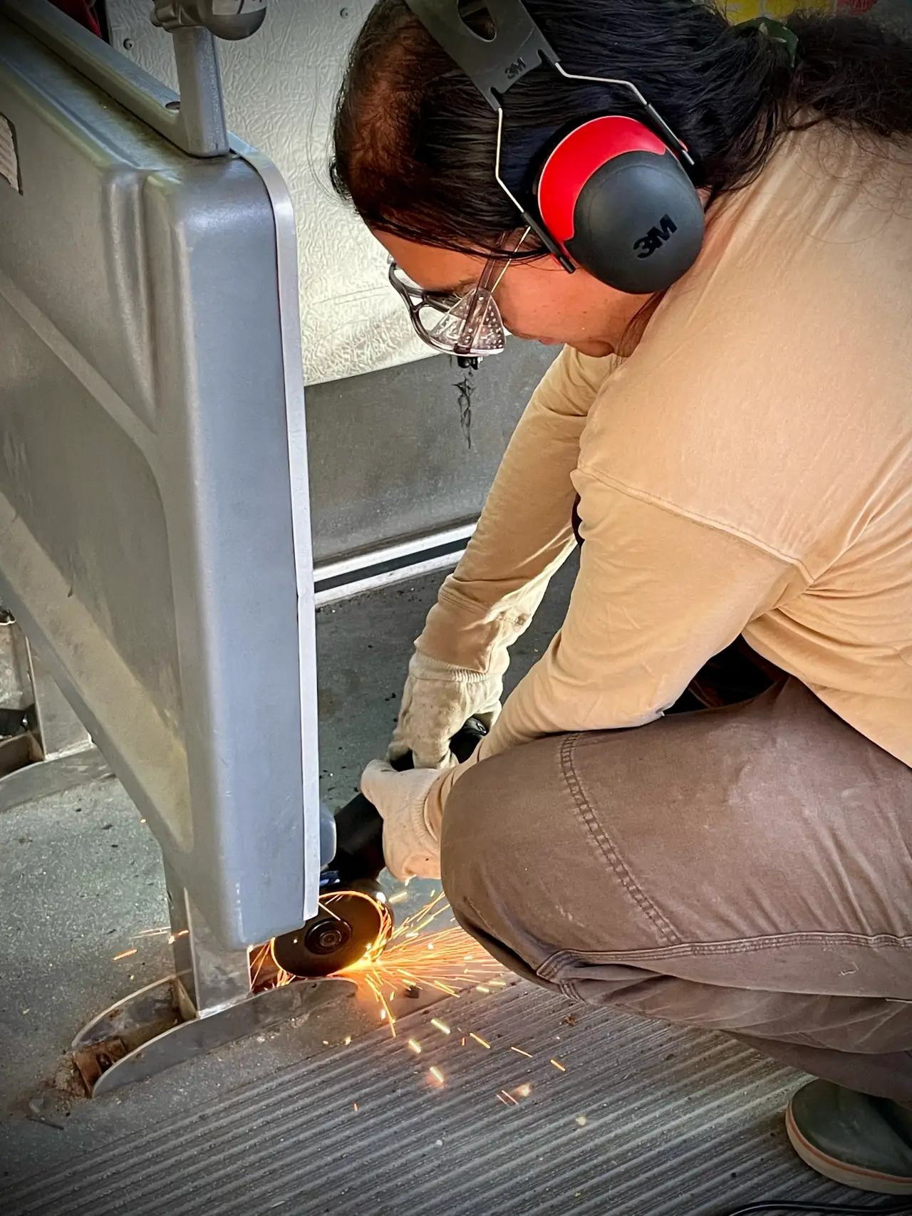 Alex kneeling on the ridged bus floor in ear defenders and safety glasses, an angle grinder throwing sparks at the foot of a grey seat frame